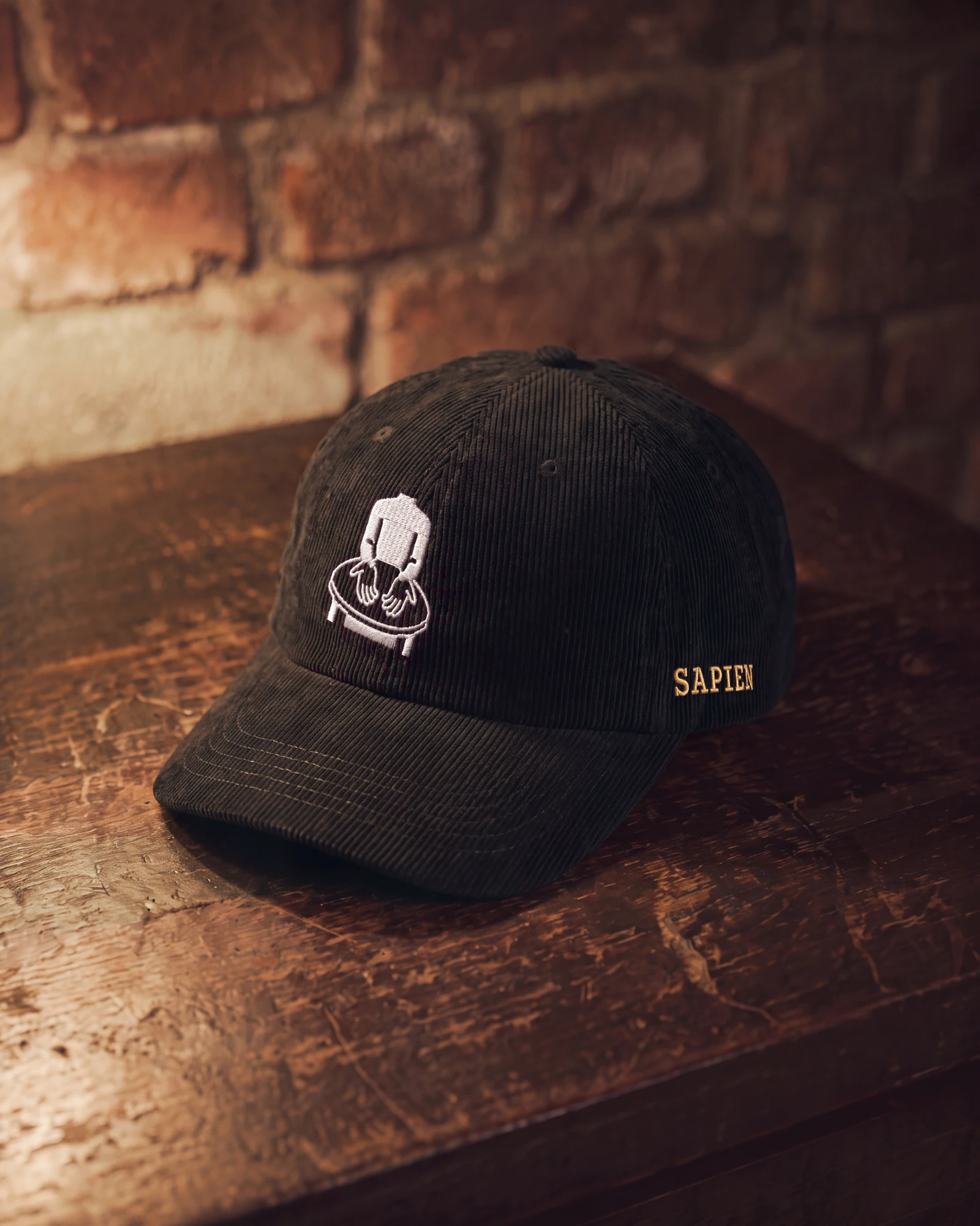 A black corduroy cap on a wooden table, the Sapien mark stitched on the front in white and SAPIEN on the left side in old gold