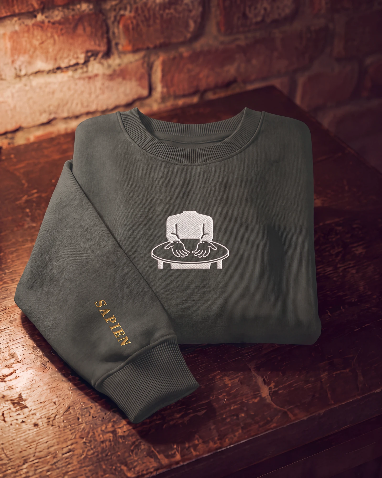 A charcoal crewneck folded on a wooden table, the Sapien mark stitched on the chest in white and SAPIEN in old gold along the sleeve