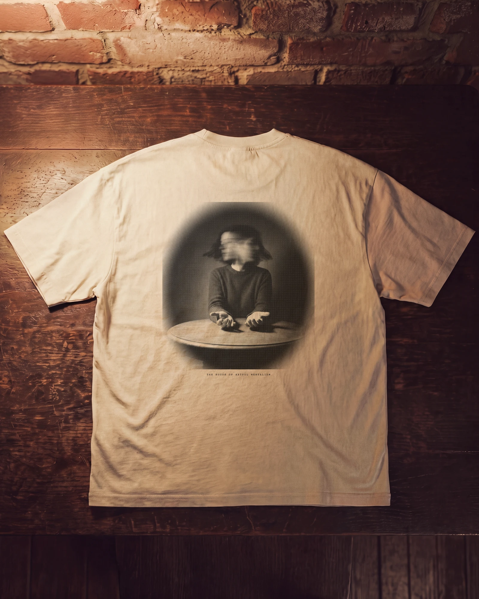 The back of a bone colored t-shirt on a wooden table, printed with a photograph of a person at a round table holding out open hands, their face lost in a blur