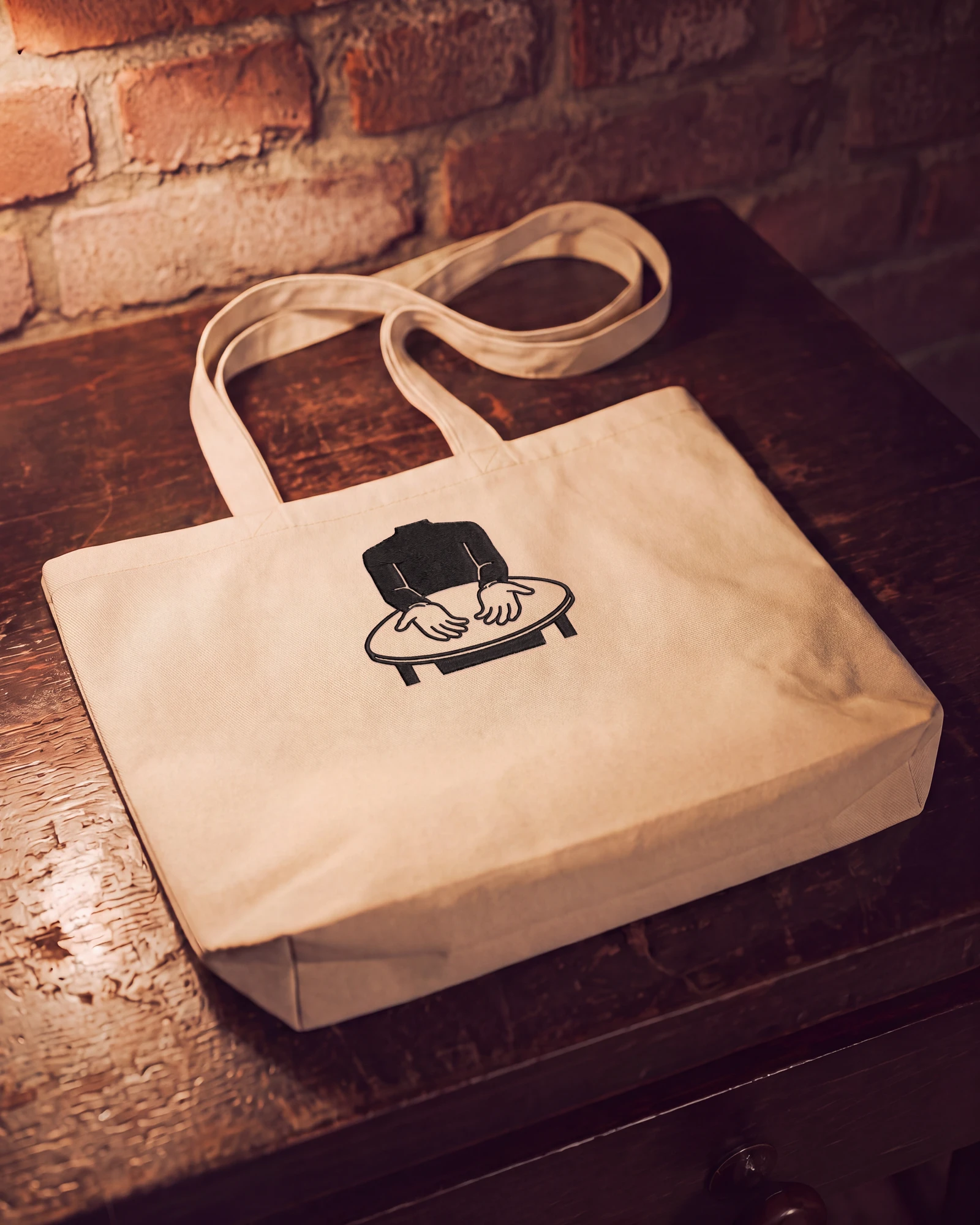 A wide oyster colored cotton tote lying on a wooden table, the Sapien mark stitched on it in black thread